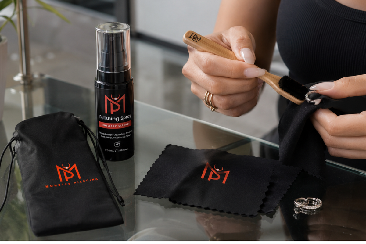 Two feminine hands clean piercing jewellery on a glass table with Monster Piercing polishing spray, black jewellery pouch, polishing cloth, bamboo brush and silver piercings.