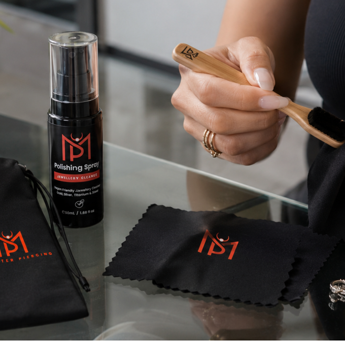 Two feminine hands clean piercing jewellery on a glass table with Monster Piercing polishing spray, black jewellery pouch, polishing cloth, bamboo brush and silver piercings.